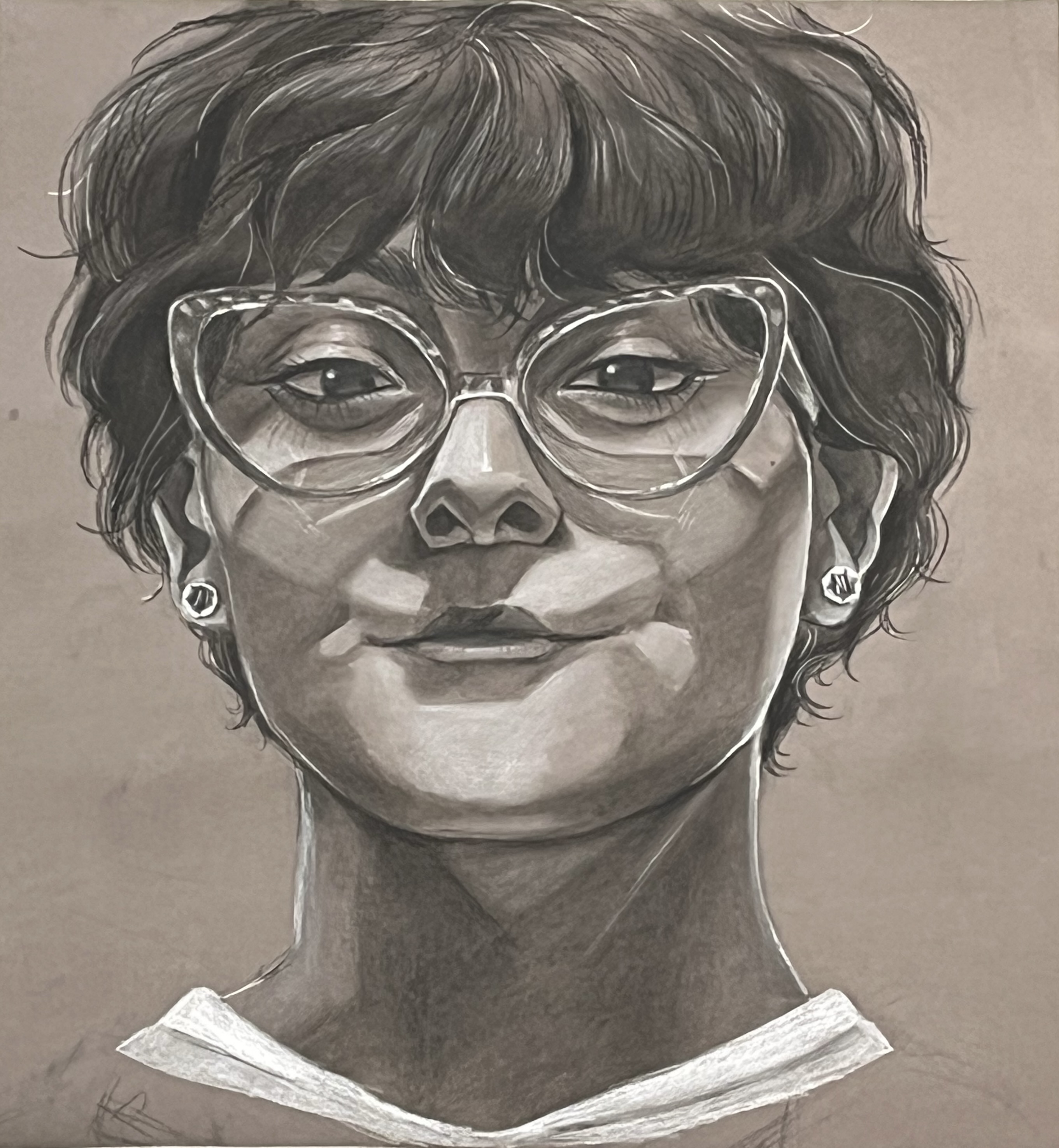 Black-and-white portrait drawing displayed in the Allan Hancock College Fine Arts Student Show. The artwork depicts a person with short, wavy hair wearing large, clear-framed glasses and small earrings. Rendered with detailed shading and soft highlights, the portrait captures a calm expression and strong facial features against a simple background.